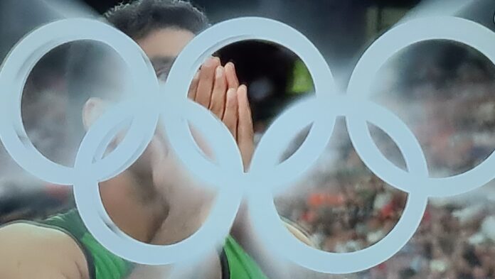 20240808_204513 A close-up photo of an athlete Arshad Nadeem with their hands covering their face, with the iconic Olympic rings blurred in the foreground. The image captures a moment of intense emotion during the Paris 2024 Olympics