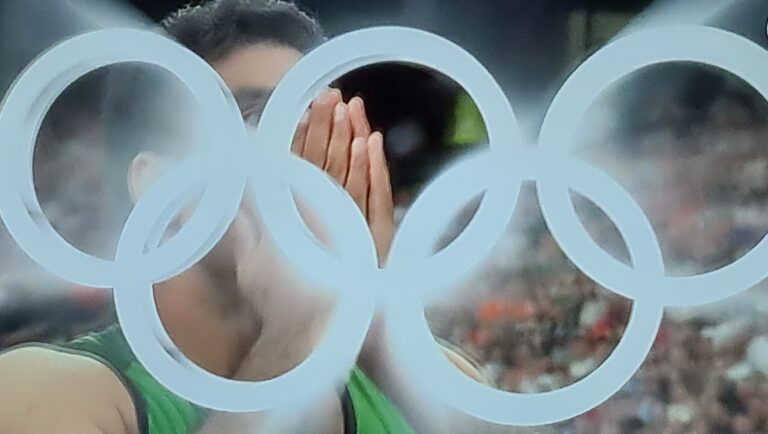 A close-up photo of an athlete Arshad Nadeem with their hands covering their face, with the iconic Olympic rings blurred in the foreground. The image captures a moment of intense emotion during the Paris 2024 Olympics
